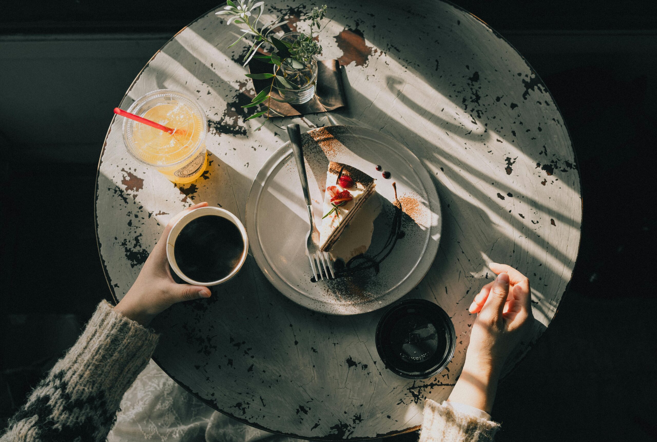 Overhead view of a cozy café table with a slice of cake, coffee, and juice, bathed in warm afternoon light.